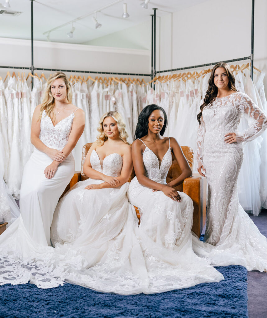 Four women modeling different white wedding gowns inside a bridal boutique showroom.