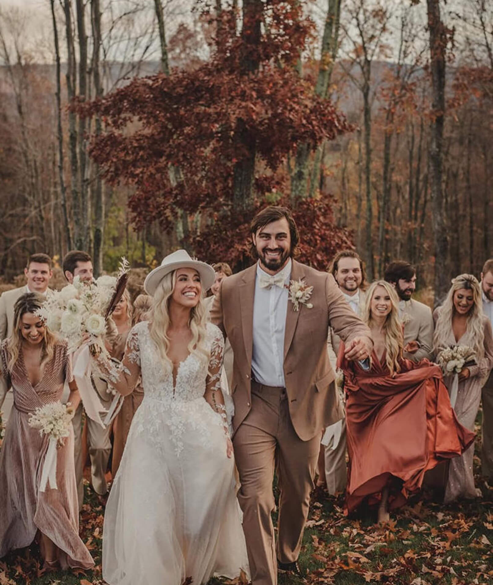 A joyful bride and groom walk with their wedding party through autumn leaves, with the bride in a long sleeve lace dress and the groom in a tan suit.