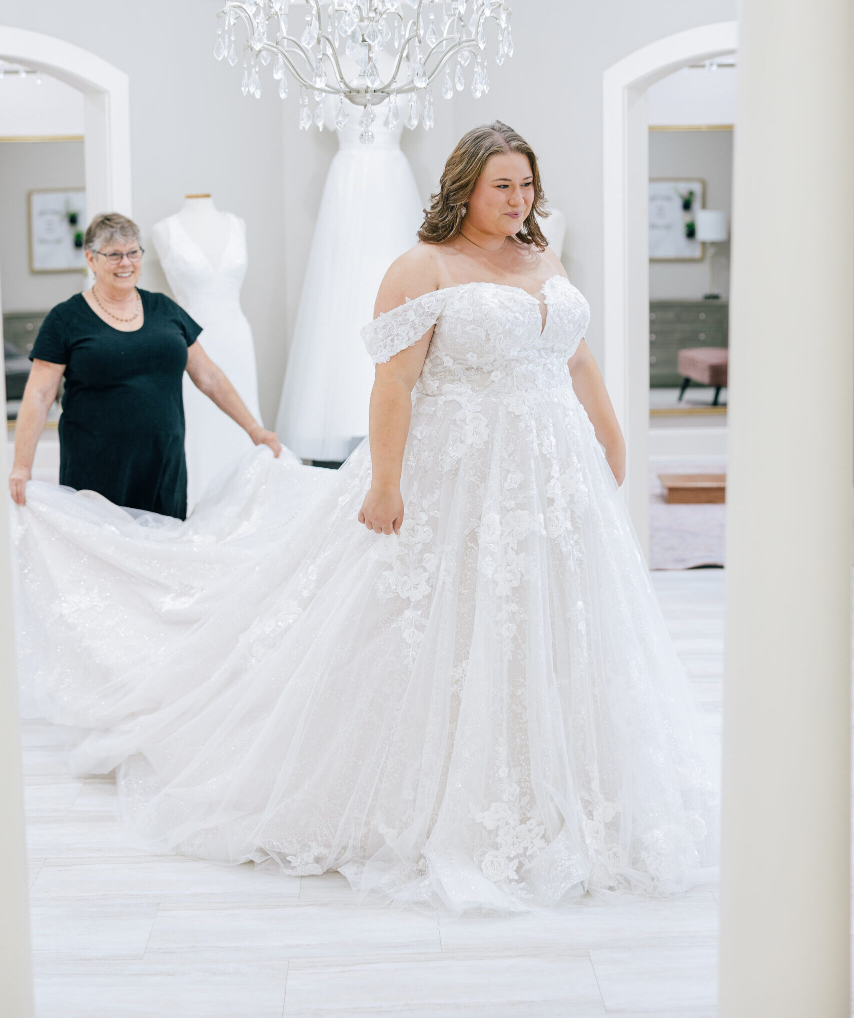 Bride stands in a sparkling lace wedding gown while an older woman holds the train behind her in a bridal salon.