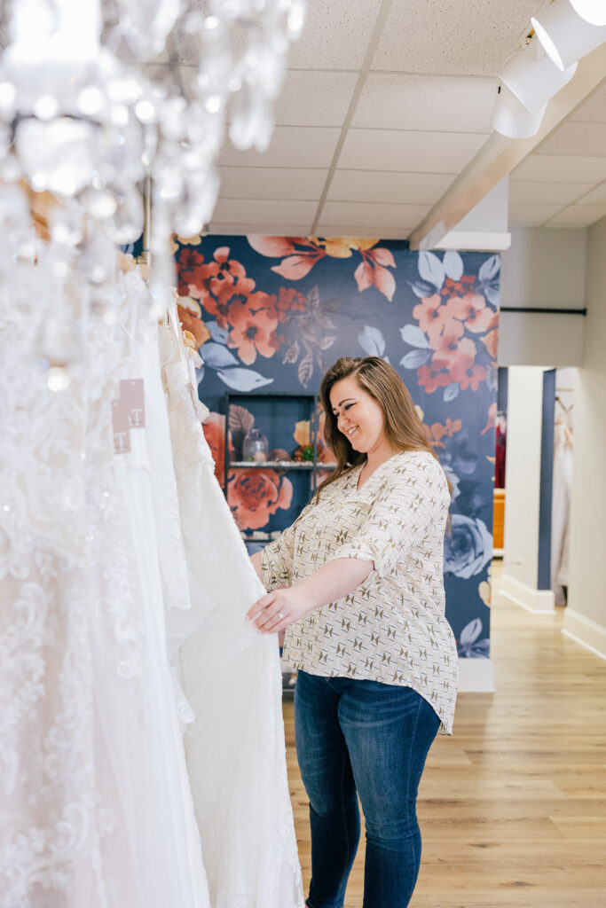 Woman browsing lace wedding gowns inside a bridal boutique with floral wallpaper in the background.