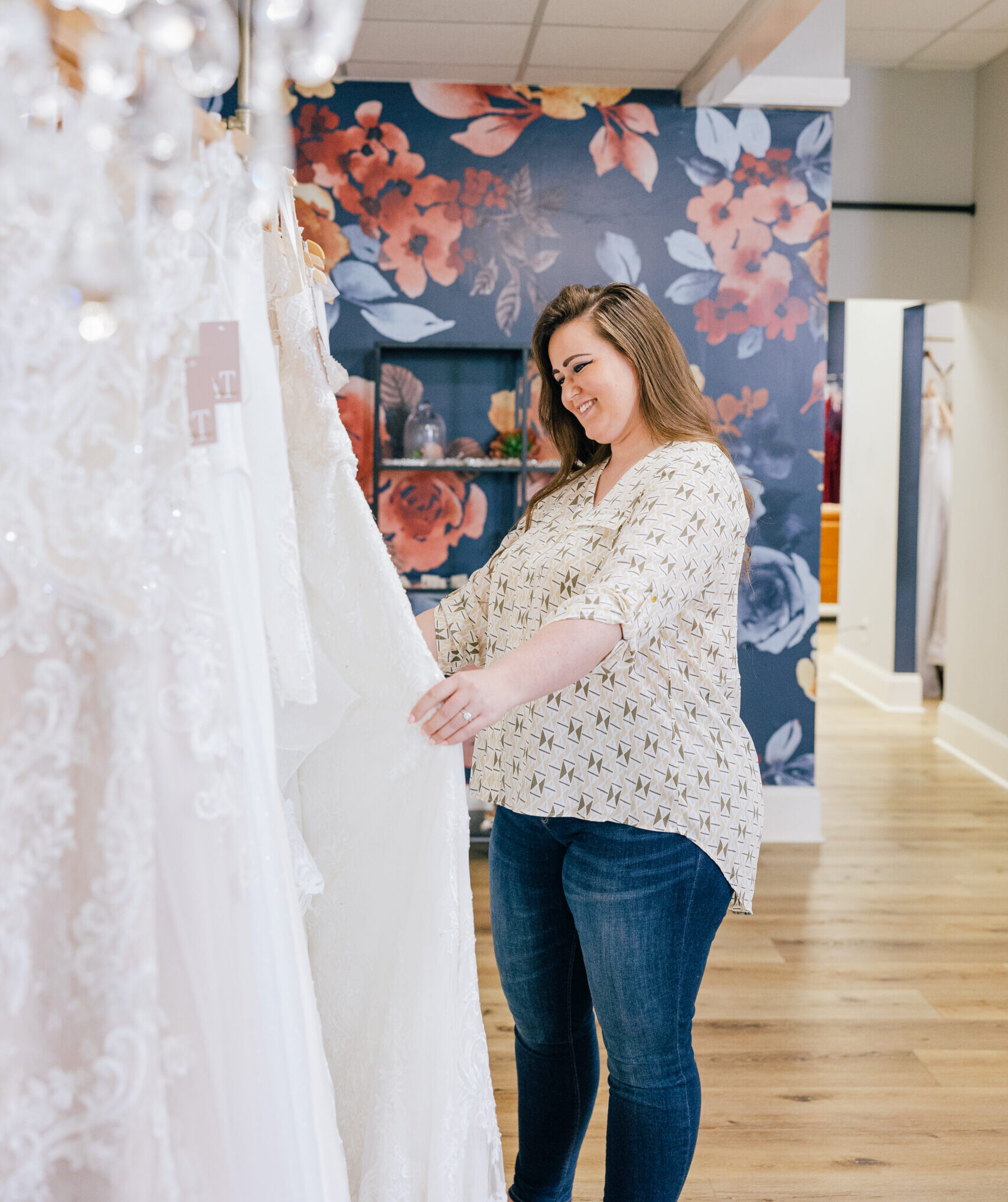 Woman browsing lace wedding gowns inside a bridal boutique with floral wallpaper in the background.