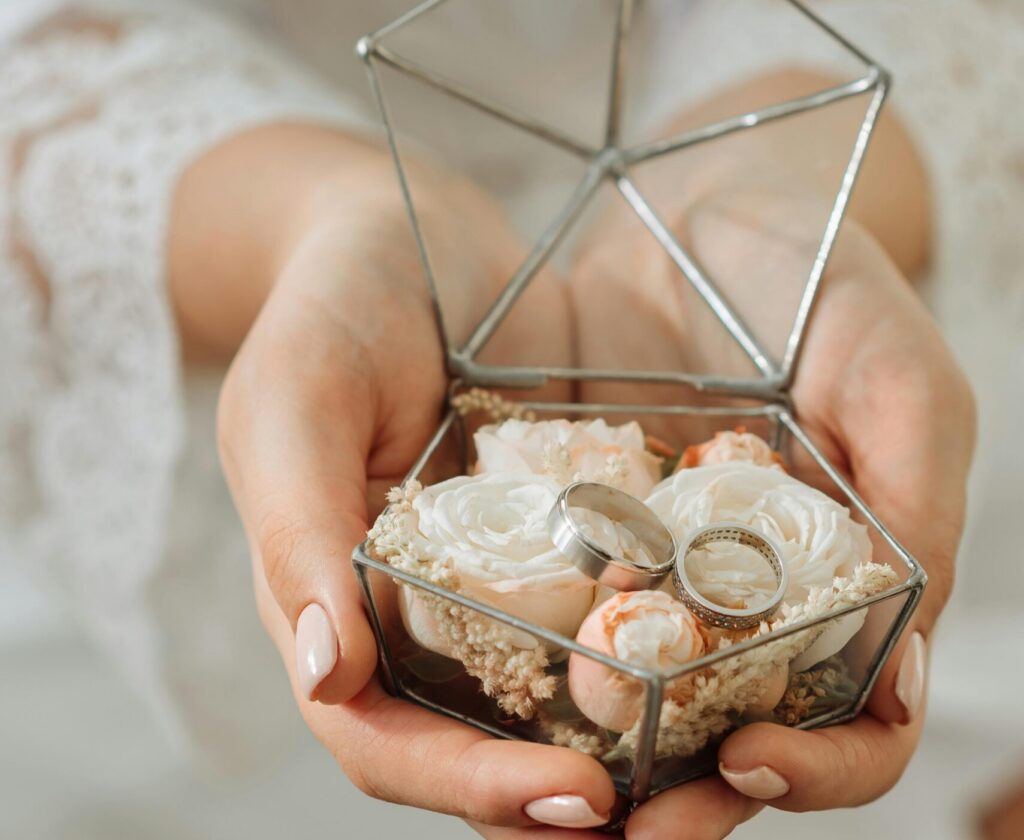 A person holds a glass ring box filled with white and blush flowers, displaying two wedding bands.