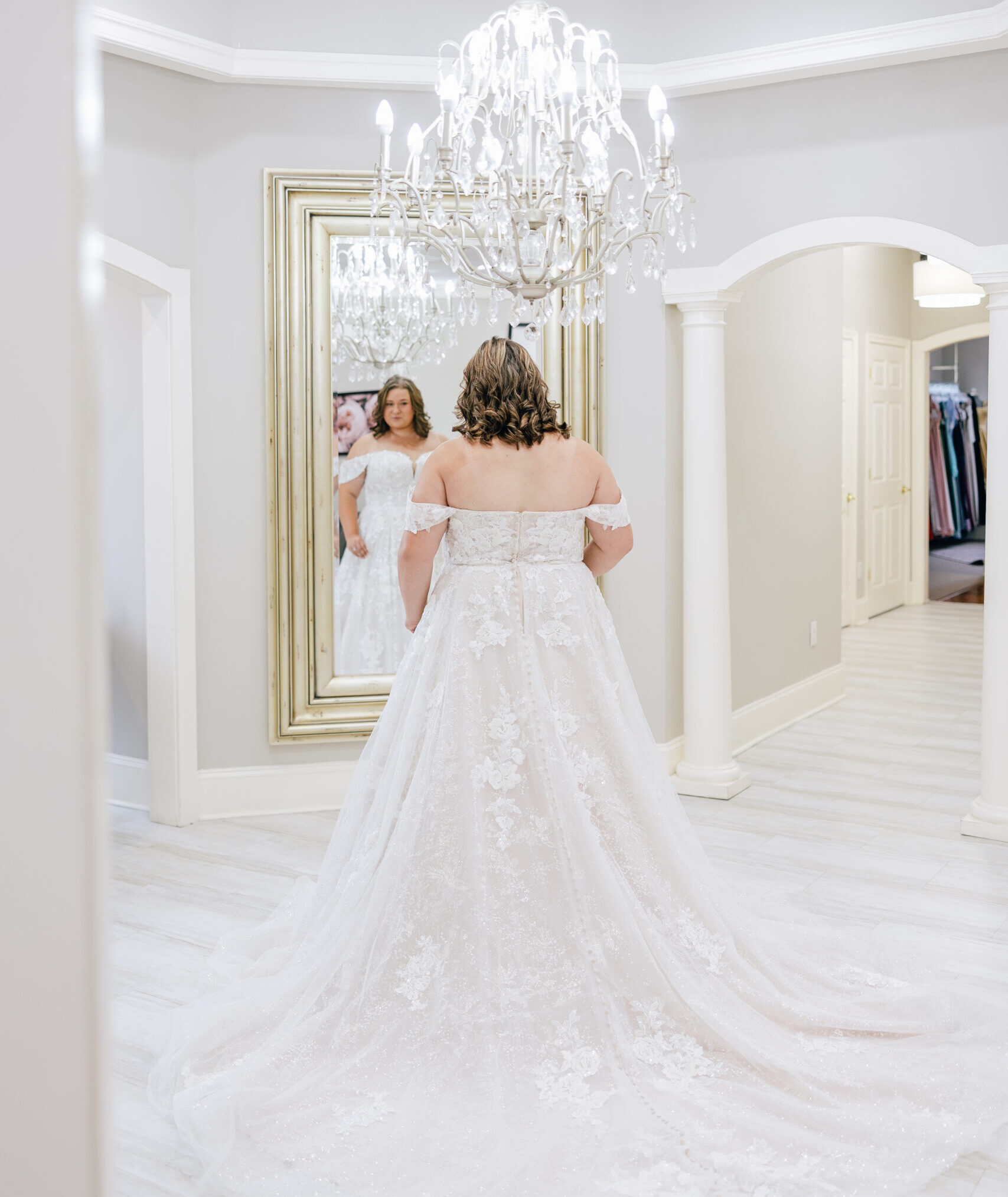 Bride views the back of her off-the-shoulder lace wedding gown while standing in front of a large mirror beneath a crystal chandelier.