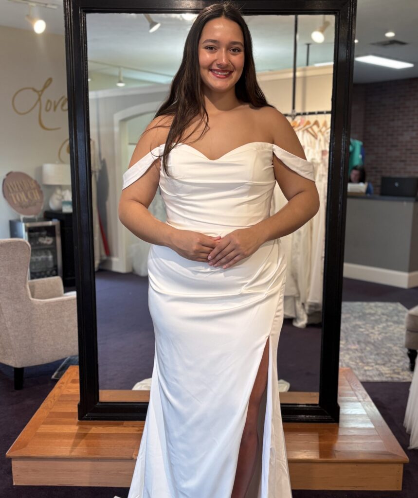 Woman wearing an off-the-shoulder satin wedding dress with a thigh-high slit stands in front of a full-length mirror in a bridal salon.