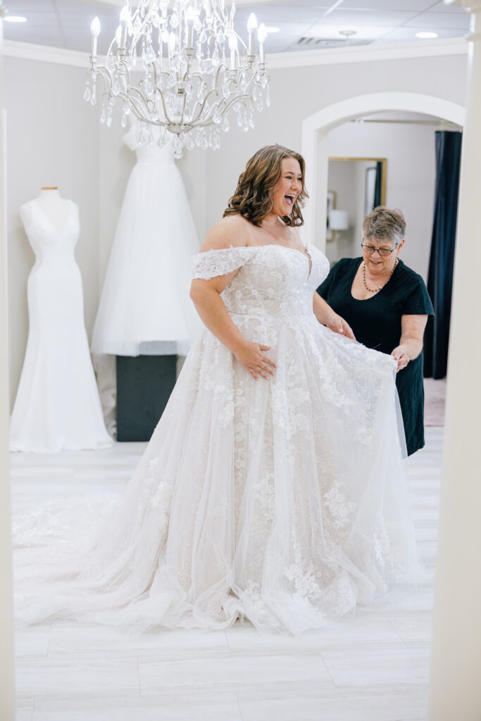 Bride smiling while trying on a lace ball gown wedding dress as another woman adjusts the skirt beneath a crystal chandelier.
