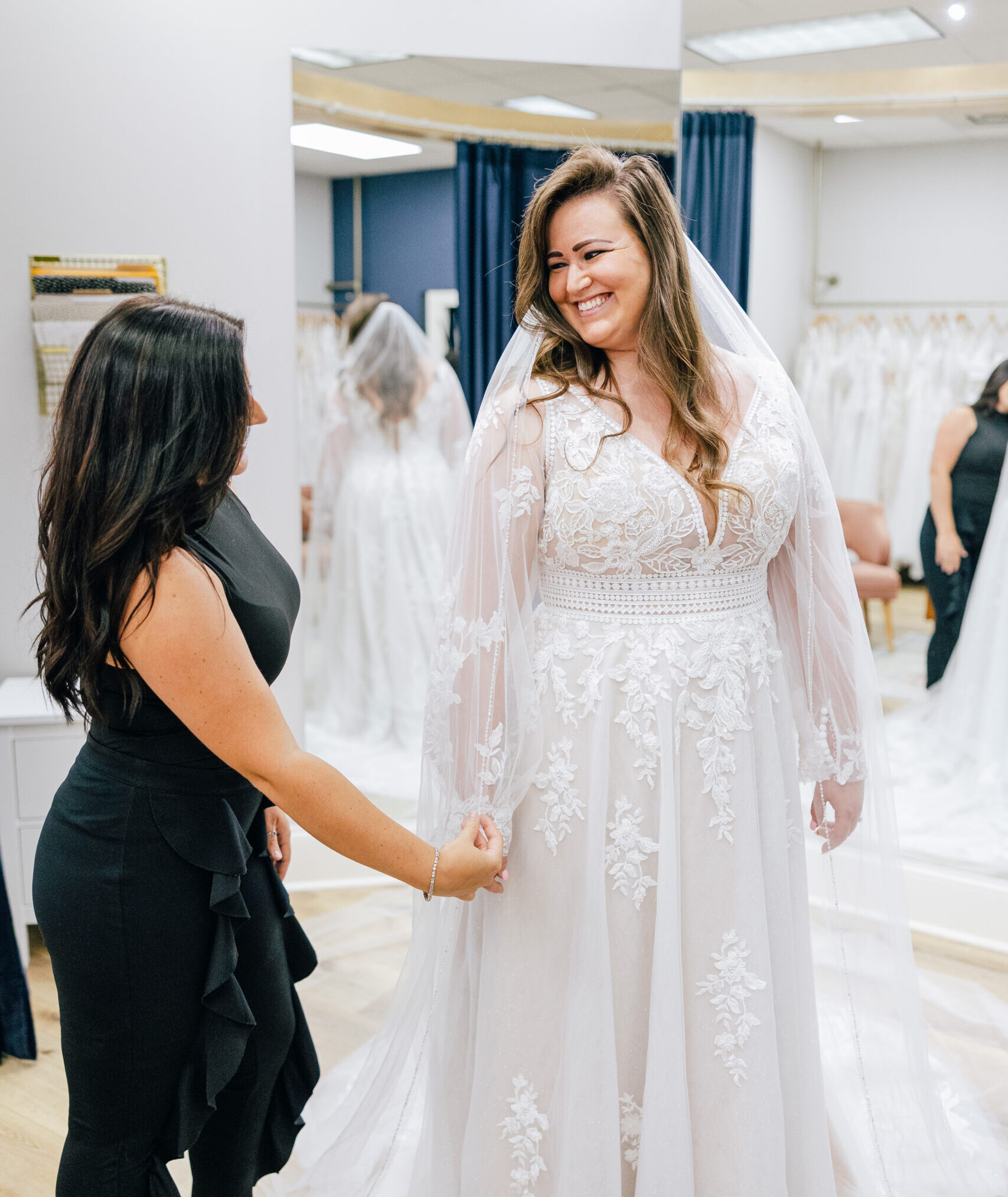 Bridal consultant adjusts a lace-edged veil as a smiling bride models a lace wedding gown in a fitting area.