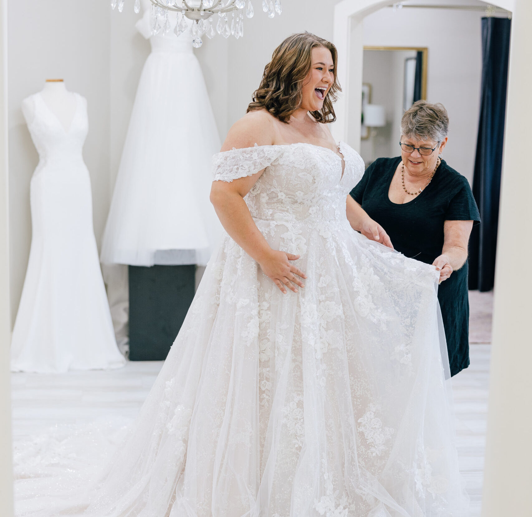 Bride smiles as an older woman adjusts the skirt of an off-the-shoulder lace wedding gown in a bright bridal boutique.