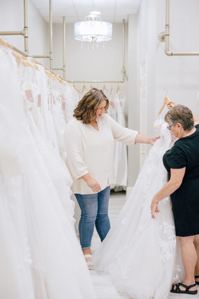 Two women examining a lace wedding gown in a bright bridal boutique lined with hanging dresses.
