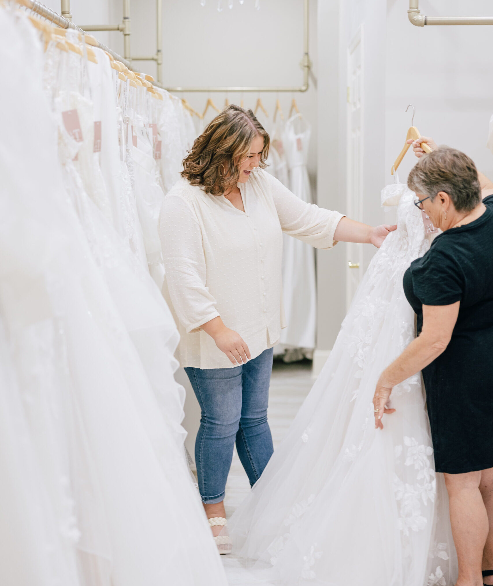 Two women examining a lace wedding gown in a bright bridal boutique lined with hanging dresses.