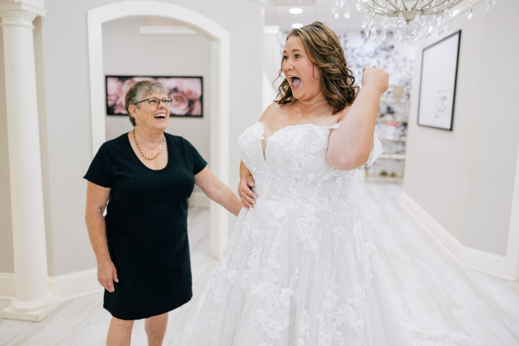 Bride reacting excitedly while wearing a floral lace wedding gown as a woman beside her smiles in a bridal salon.