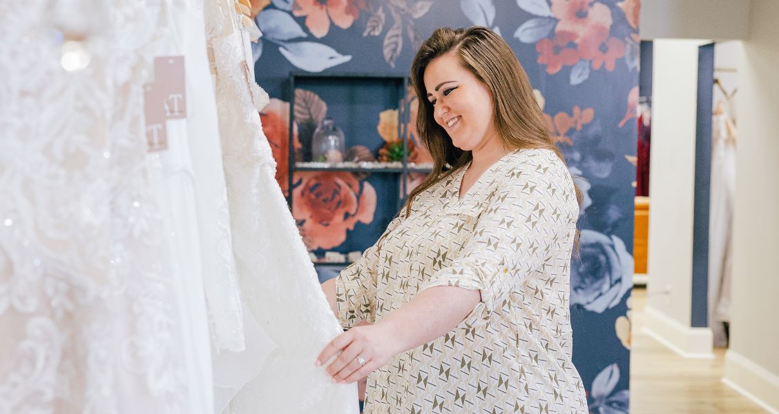 Woman smiling while looking at a wedding dress rack inside a bridal boutique with floral accent walls.