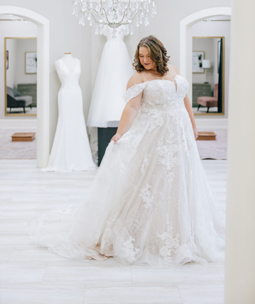 Bride wearing an off-the-shoulder lace ballgown poses beneath a crystal chandelier in an elegant bridal showroom.