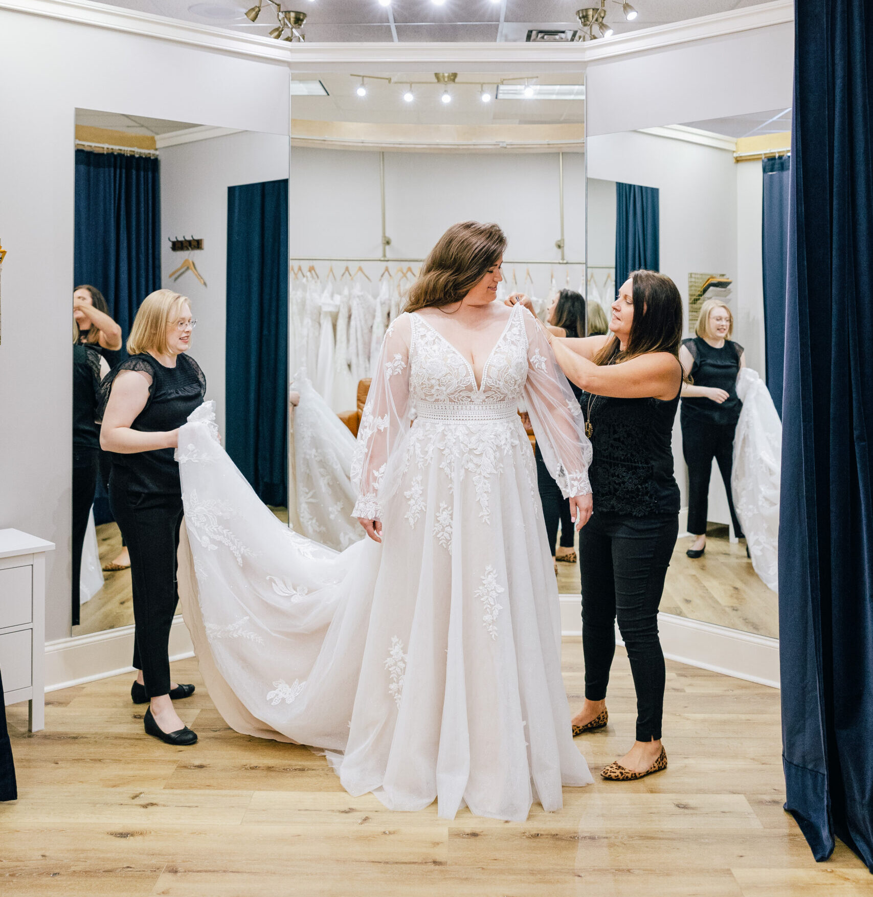 Bridal consultants adjust the sleeves and train of a lace wedding gown during a fitting appointment.
