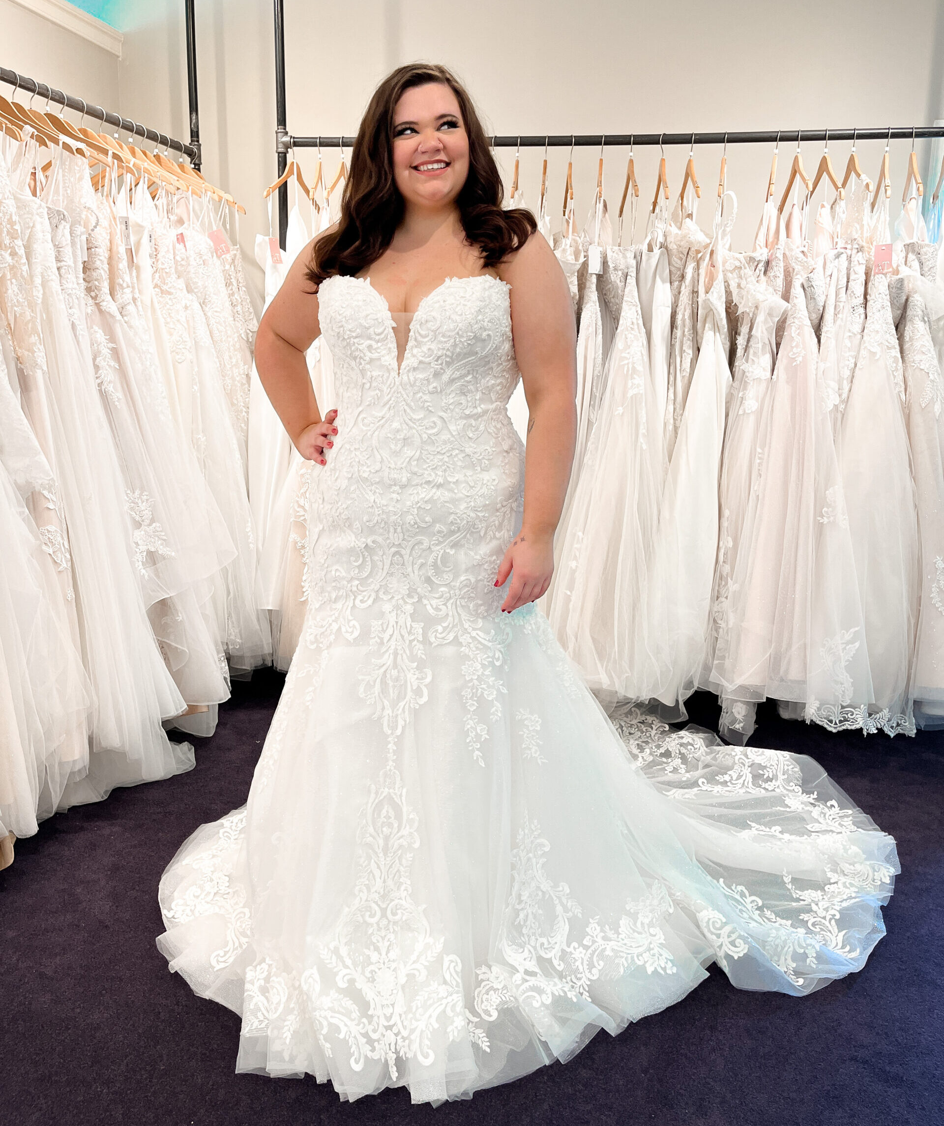 Smiling bride models a fitted lace wedding dress with a long train in a bridal boutique surrounded by wedding gowns.