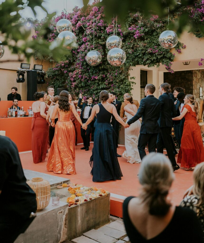 Wedding guests hold hands and dance in a circle beneath hanging disco balls and flowering greenery at an outdoor reception.