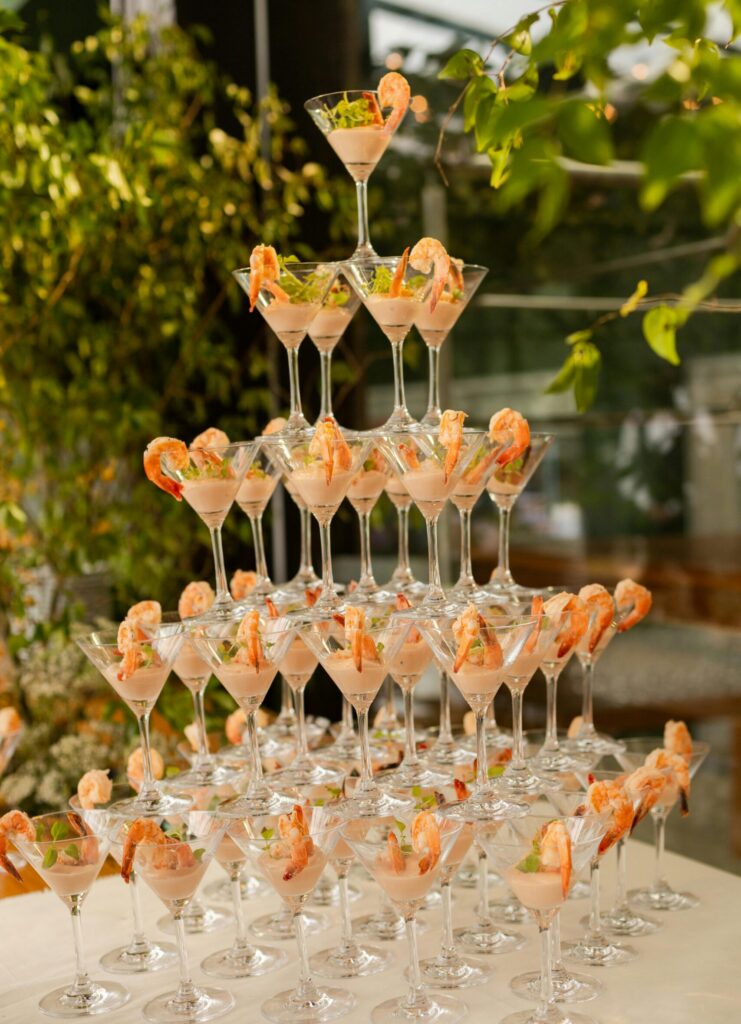 Shrimp cocktail appetizers served in martini glasses are stacked in a pyramid display surrounded by lush greenery.