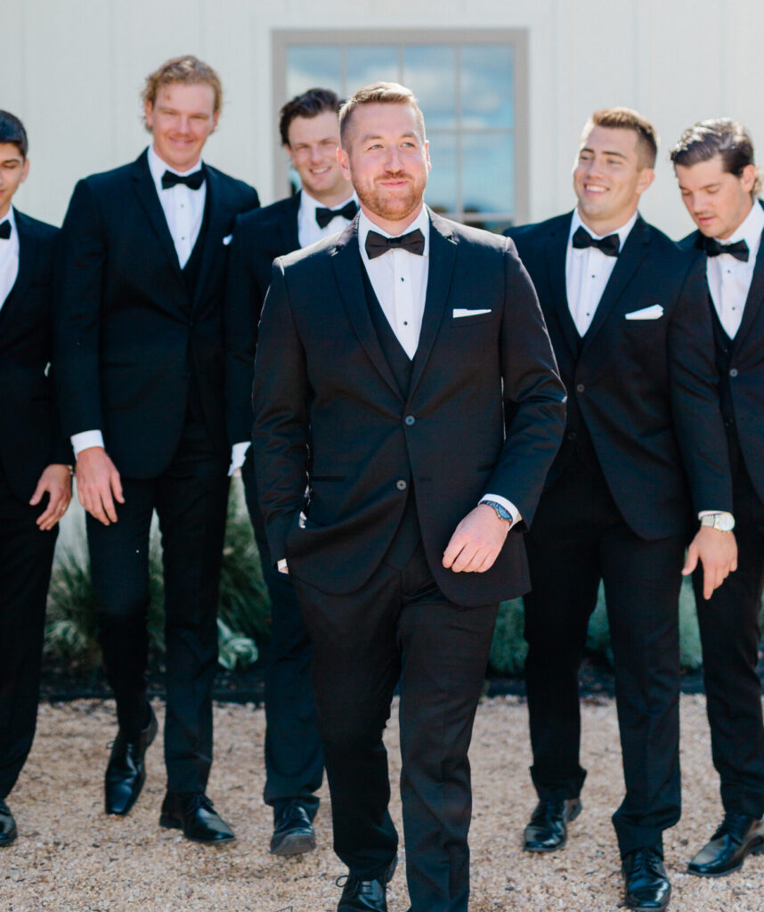A groom in a classic black tuxedo walks confidently in front of his smiling groomsmen, all dressed in black tuxedos, outside a white building.