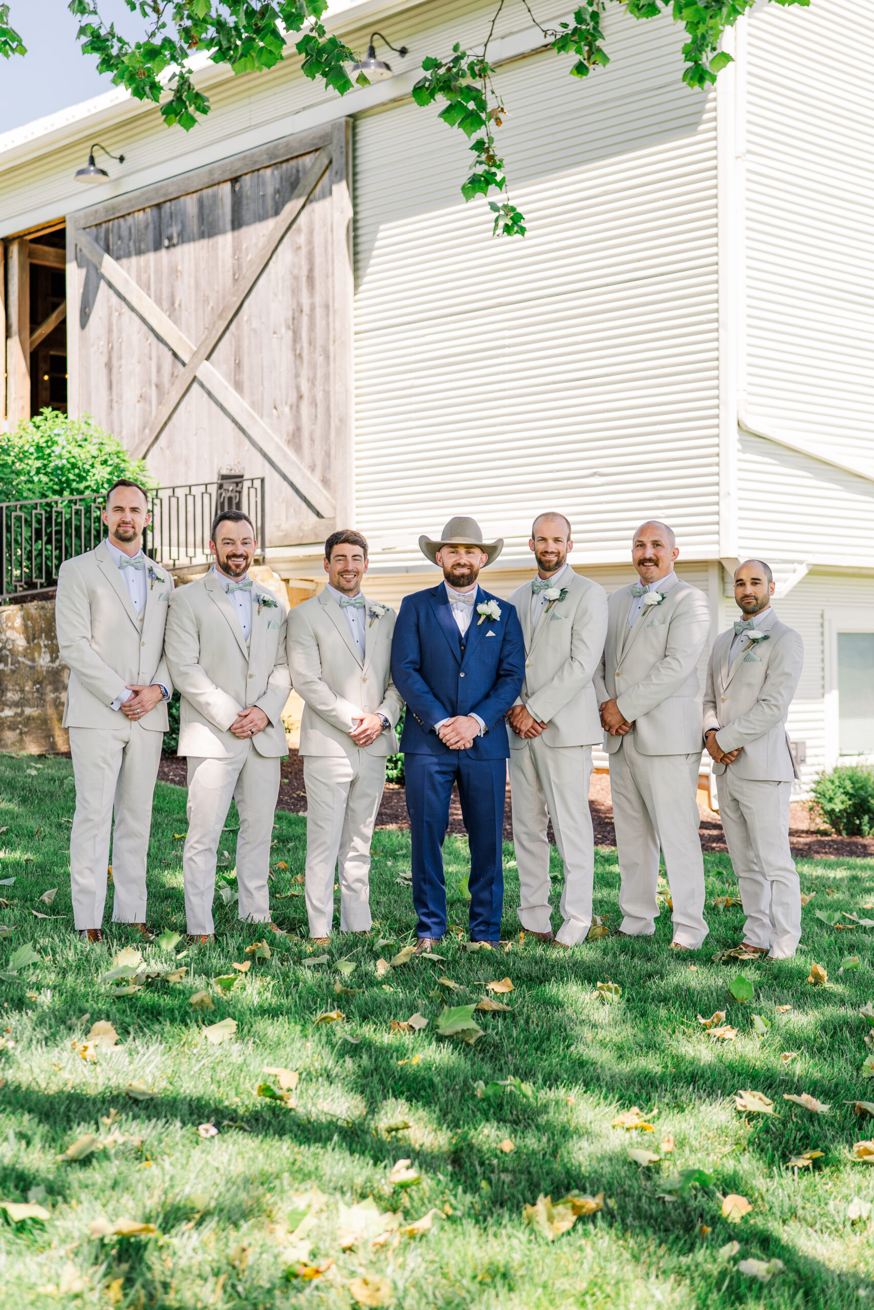 A groom in a blue suit and cowboy hat stands with six groomsmen in matching light beige suits outside a rustic barn on a sunny day.