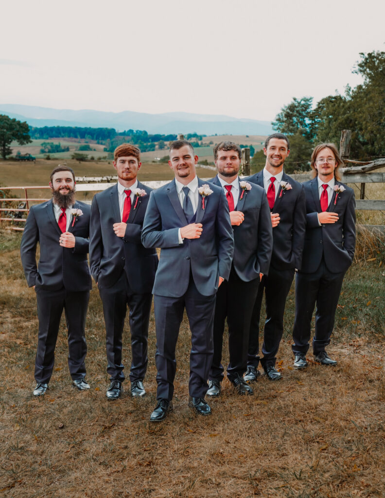 Groom and five groomsmen pose together outdoors in matching dark suits with red ties against a scenic countryside backdrop.