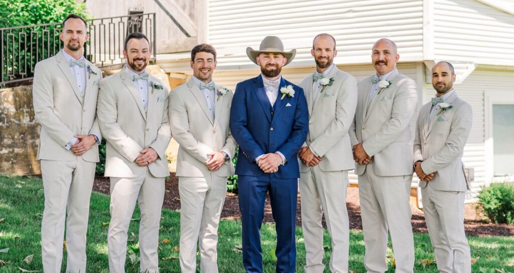 Groom in a blue suit and cowboy hat poses with six groomsmen in matching light beige suits on the lawn outside a rustic barn-style venue.