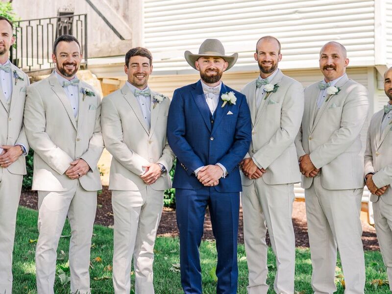 Groom in a blue suit and cowboy hat poses with six groomsmen in matching light beige suits on the lawn outside a rustic barn-style venue.