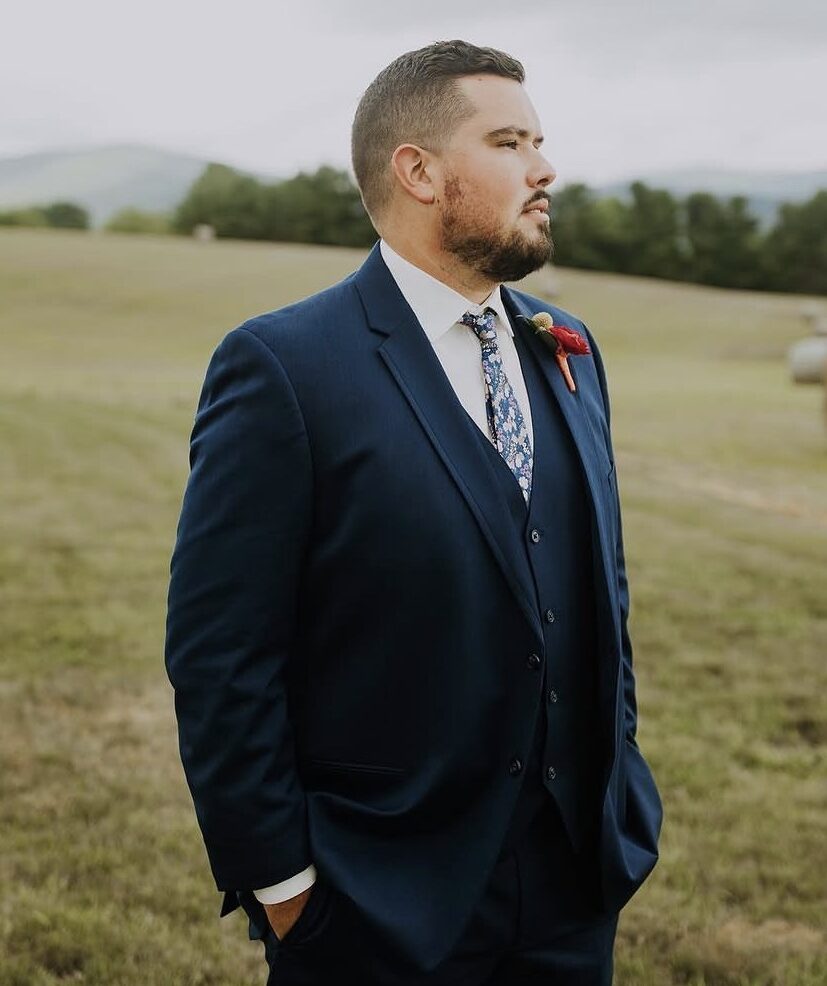 A groom wearing a navy blue three-piece suit with a floral tie stands in an open field with hay bales and distant hills behind him.