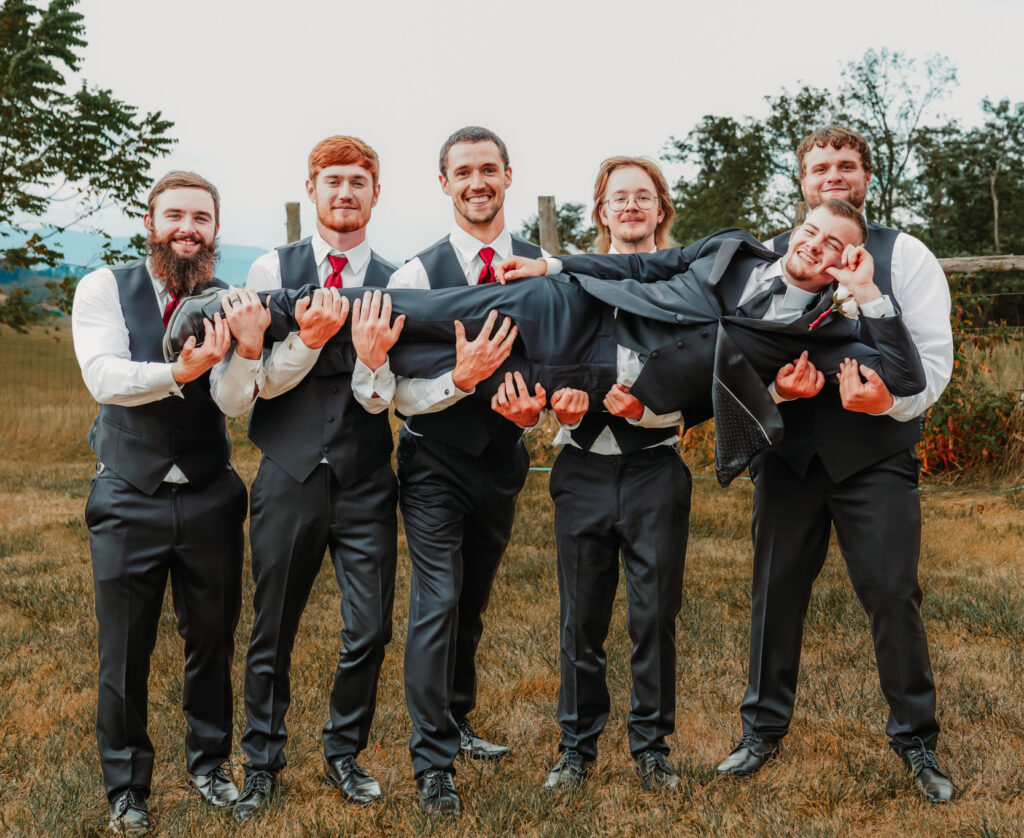 Six groomsmen wearing matching dark gray vests, white shirts, and red ties hold the groom horizontally while he smiles and poses during an outdoor wedding portrait.
