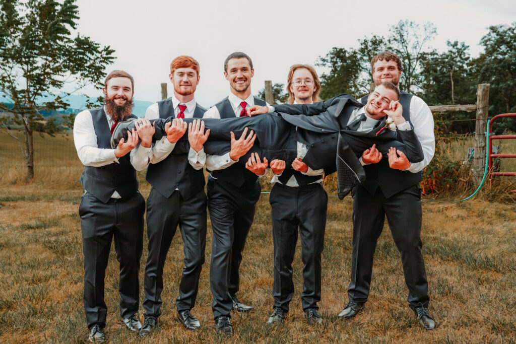 Six groomsmen wearing matching dark gray vests, white shirts, and red ties hold the groom horizontally while he smiles and poses during an outdoor wedding portrait.
