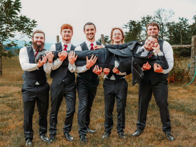 Six groomsmen wearing matching dark gray vests, white shirts, and red ties hold the groom horizontally while he smiles and poses during an outdoor wedding portrait.