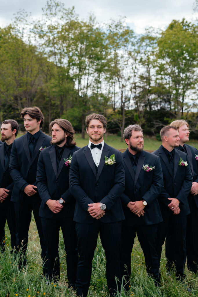 A groom in a black tuxedo stands in front of his groomsmen, who are dressed in coordinated black suits and boutonnieres, in a grassy field with trees in the background.