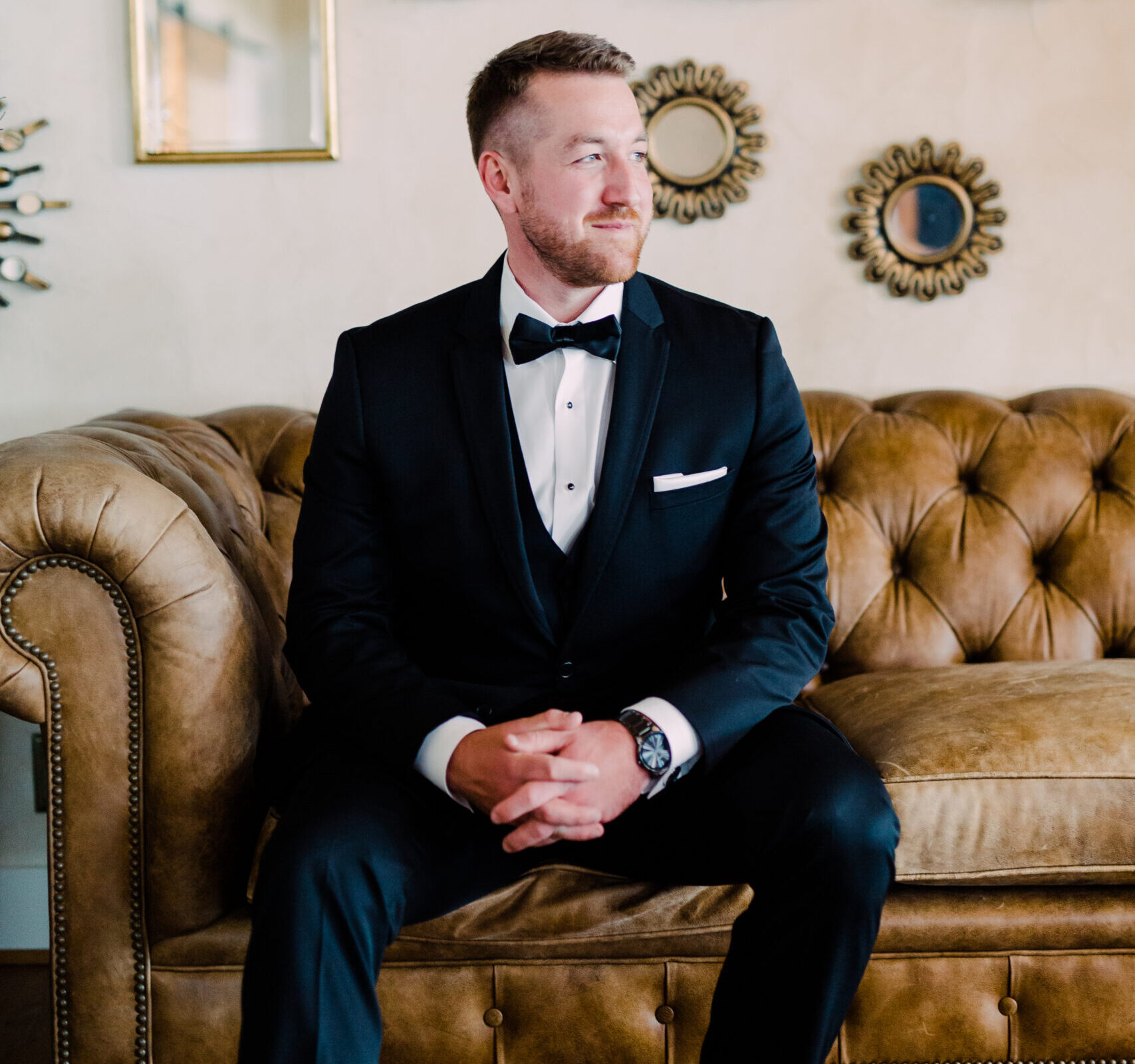 A groom wearing a classic black tuxedo sits on a tufted leather sofa beneath ornate gold-framed mirrors in an elegant room.