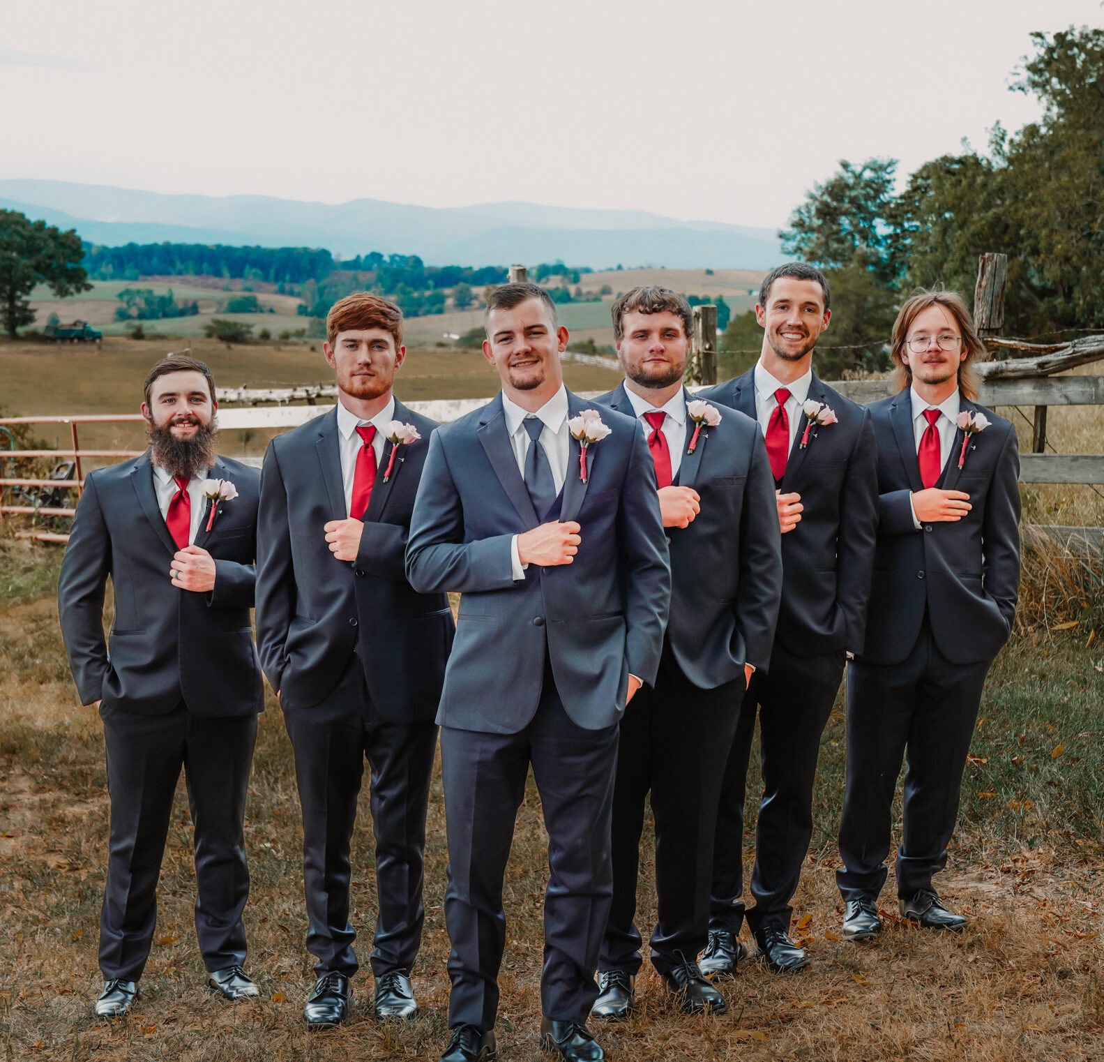 A groom in a blue suit stands in front of six groomsmen wearing dark suits with red ties, posing outdoors with rolling hills and a rustic fence in the background.
