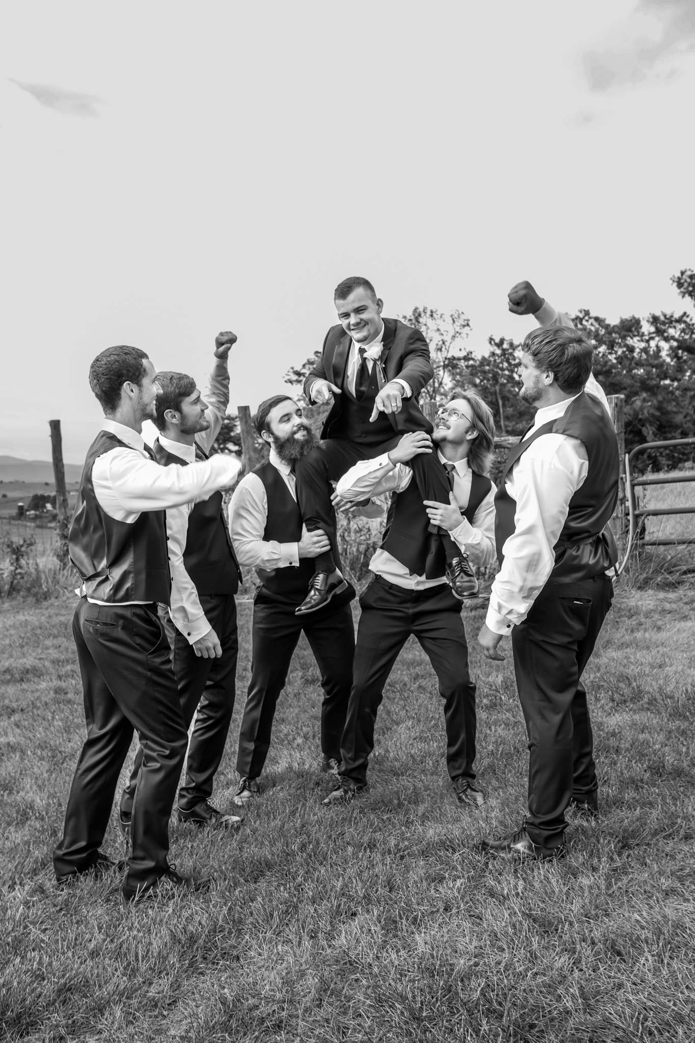 Six groomsmen lift the groom onto their shoulders while celebrating together in an open field during an outdoor wedding.