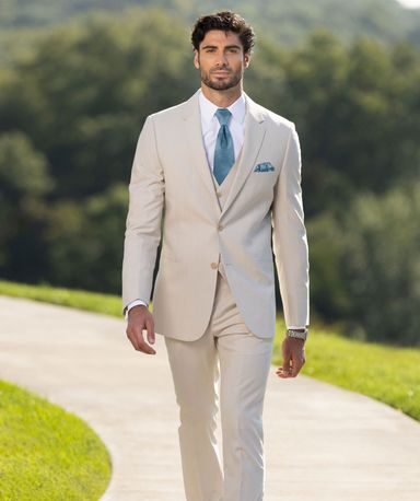 A man wearing a light cream suit with a blue tie walks confidently along a paved path surrounded by green landscaping.