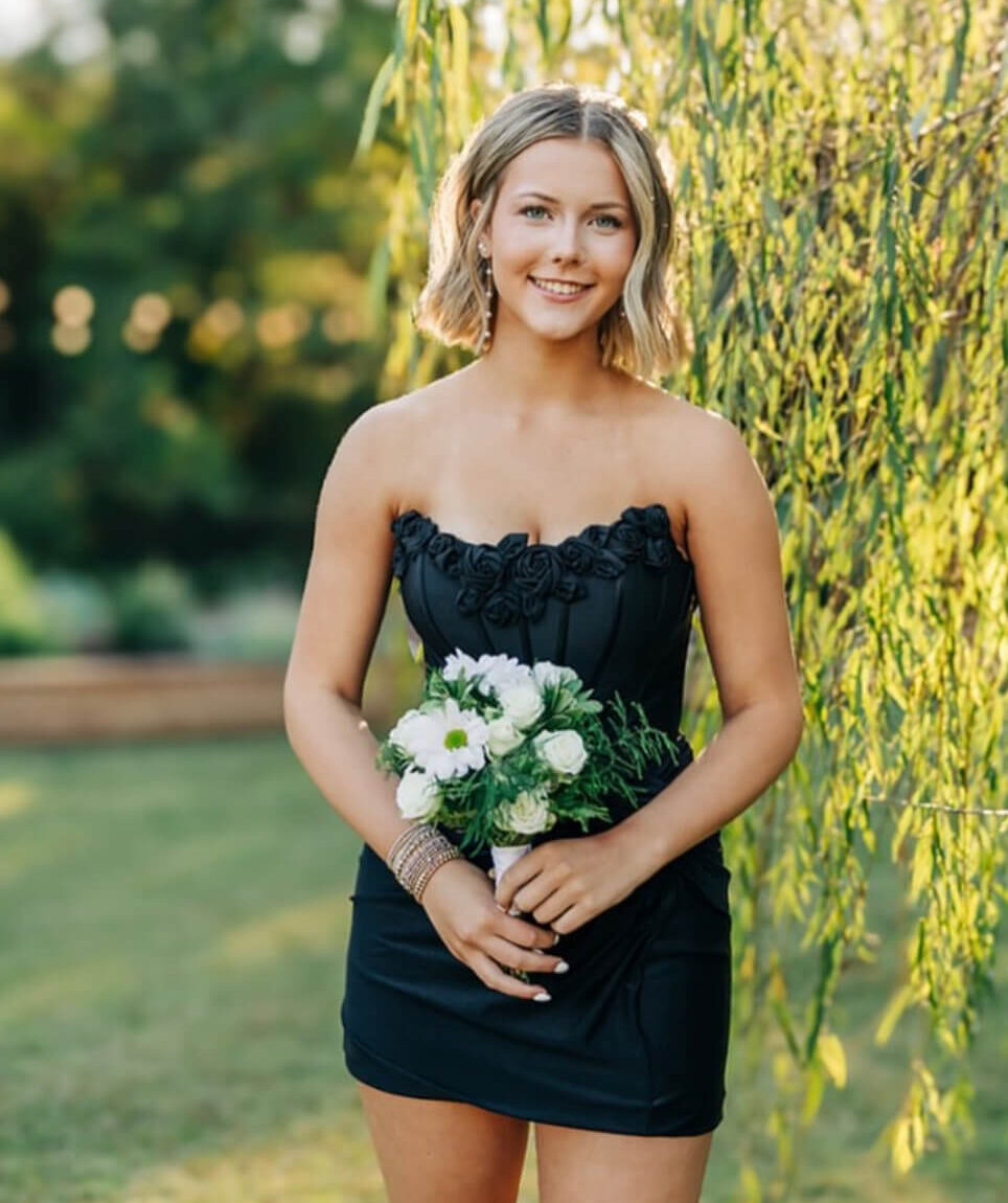 A woman poses outdoors holding a white floral bouquet while wearing a short black strapless dress with floral details along the neckline.
