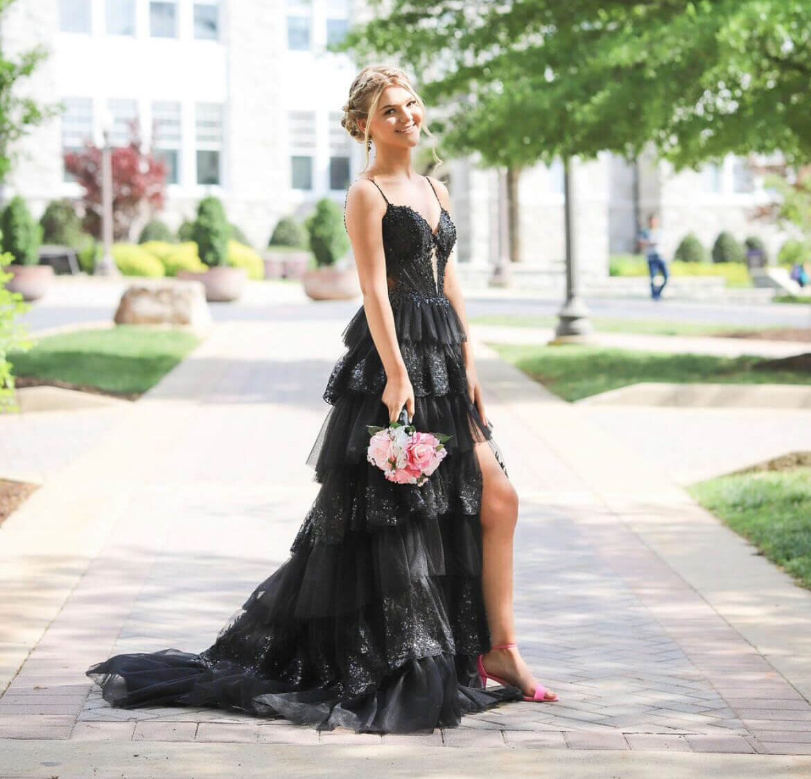 A woman poses outdoors in a dramatic black tiered gown with a high slit while holding a bouquet of pink flowers.