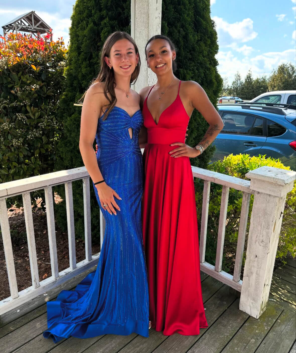 Two women pose together on a porch, one wearing a sparkling blue floor-length gown and the other wearing a red floor-length gown.