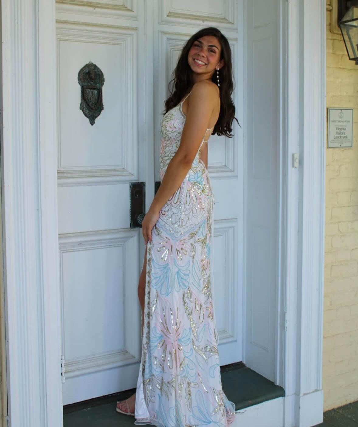 A woman poses in front of large white doors wearing a floor-length white gown with pastel floral patterns and shimmering embellishments.