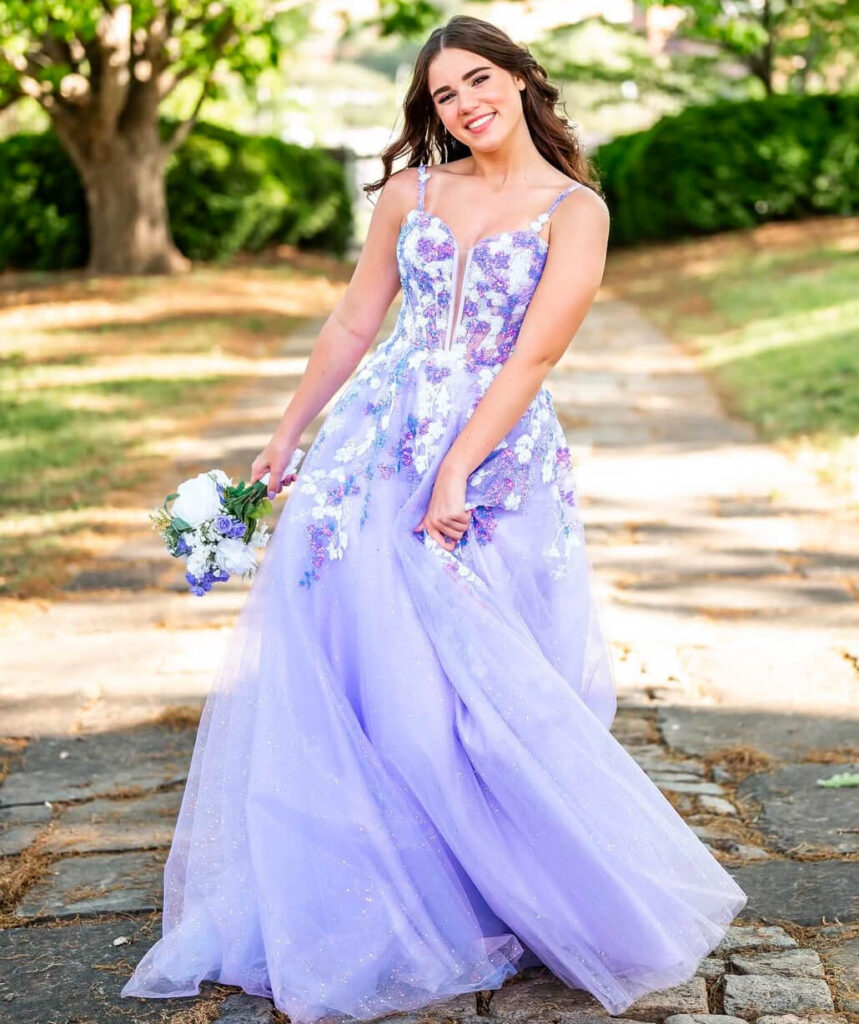 A woman poses outdoors in a floor-length lavender gown with a floral embellished bodice while holding a small bouquet.x