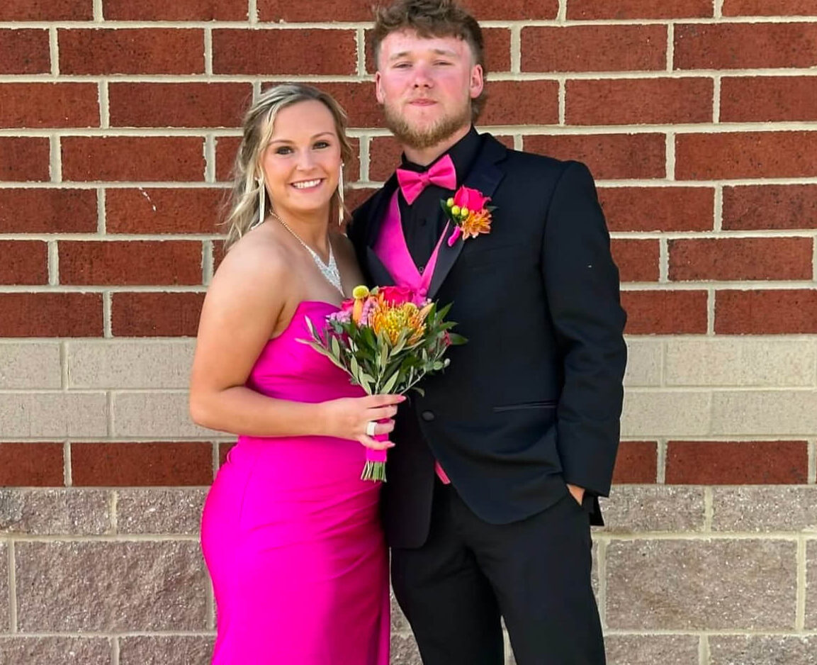 A couple poses in front of a brick wall, with the woman wearing a bright pink strapless floor-length gown and holding a colorful bouquet and the man wearing a black suit with pink accessories.