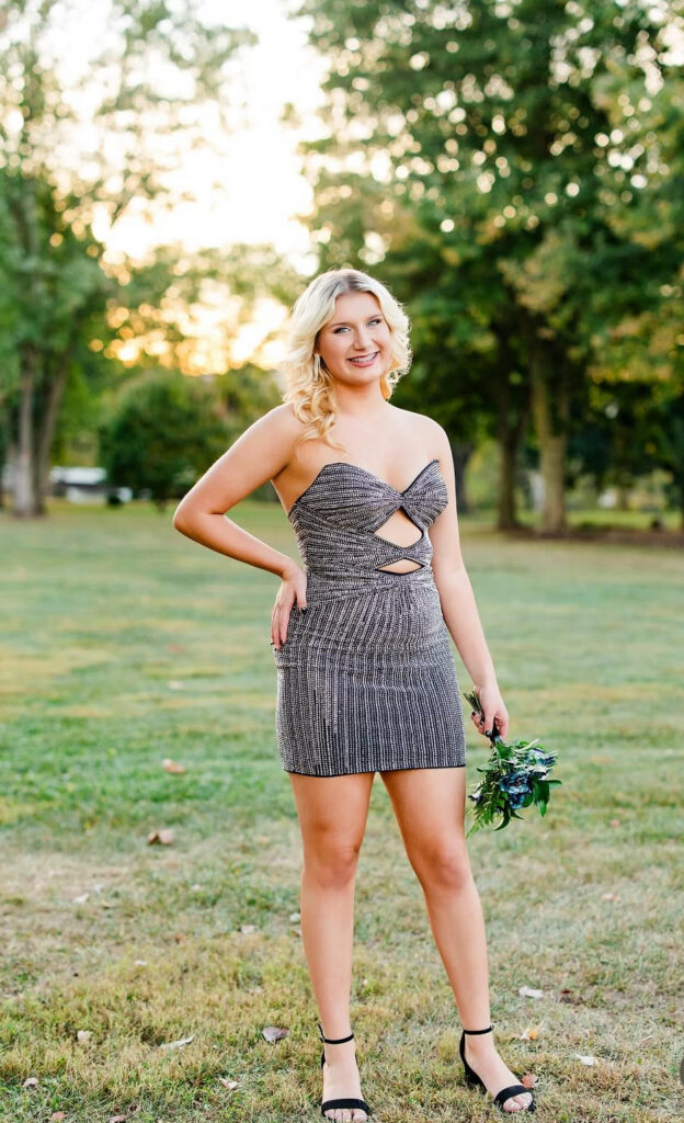A woman poses outdoors in a sparkling silver mini dress with a strapless twisted bodice, front cutouts, and black ankle-strap heels.
