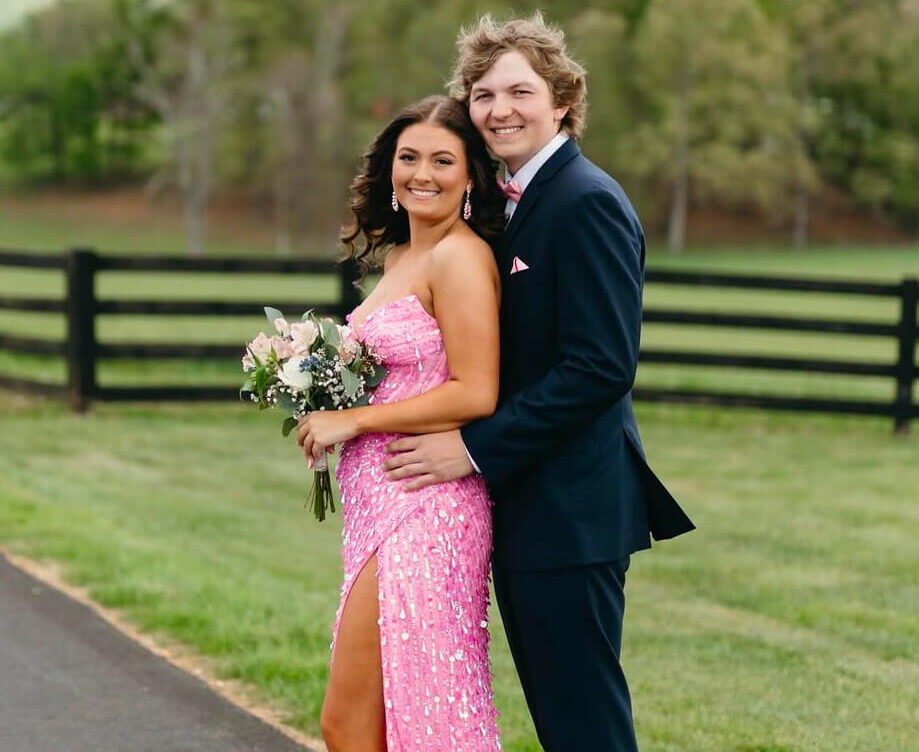 A couple poses outdoors near a black fence, with the woman wearing a sparkling pink strapless gown with a high slit and holding a bouquet and the man wearing a navy suit with pink accessories.