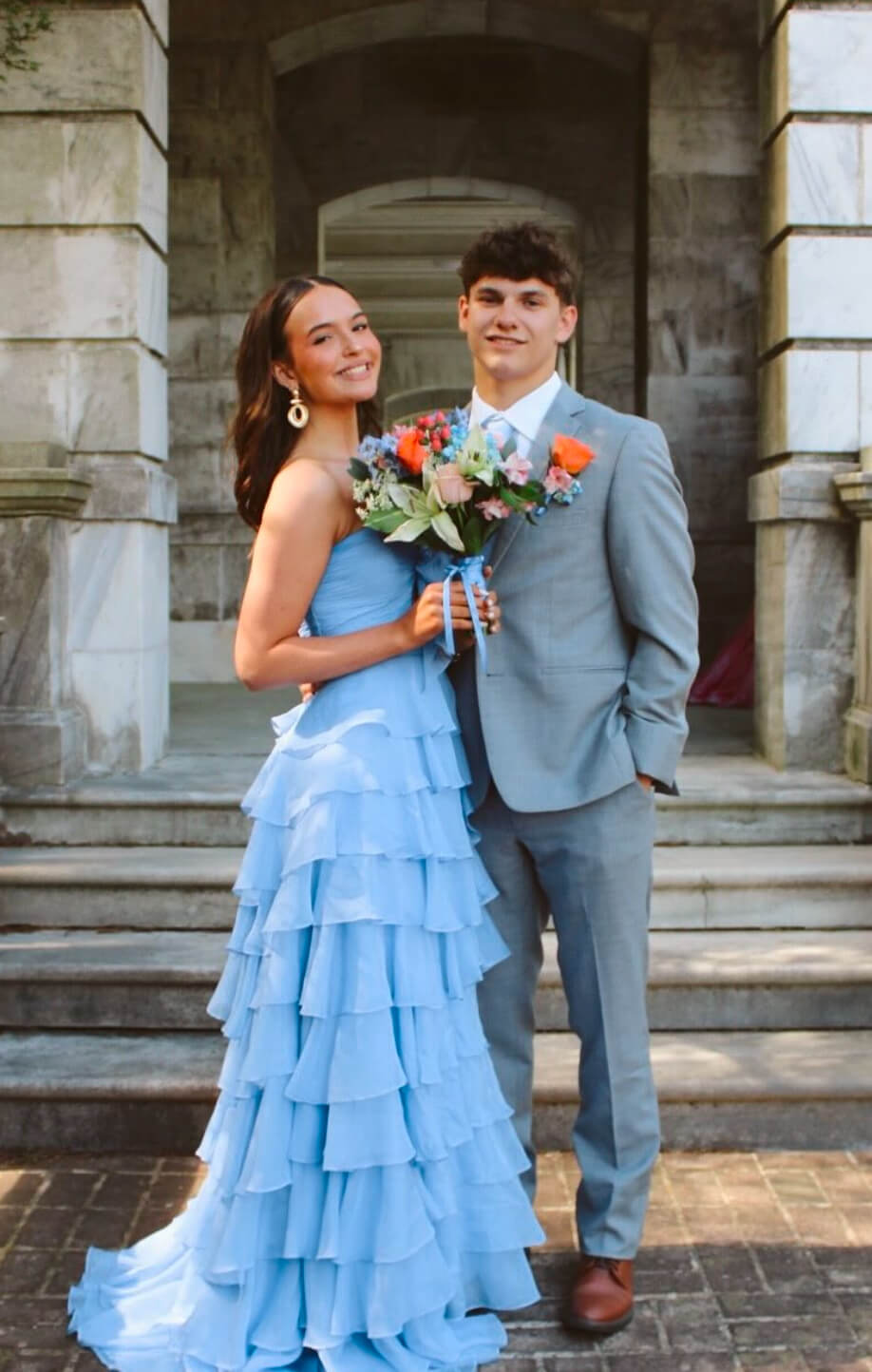 A couple poses on the steps of a stone building, with the woman wearing a light blue strapless tiered gown and holding a colorful bouquet and the man wearing a gray suit.