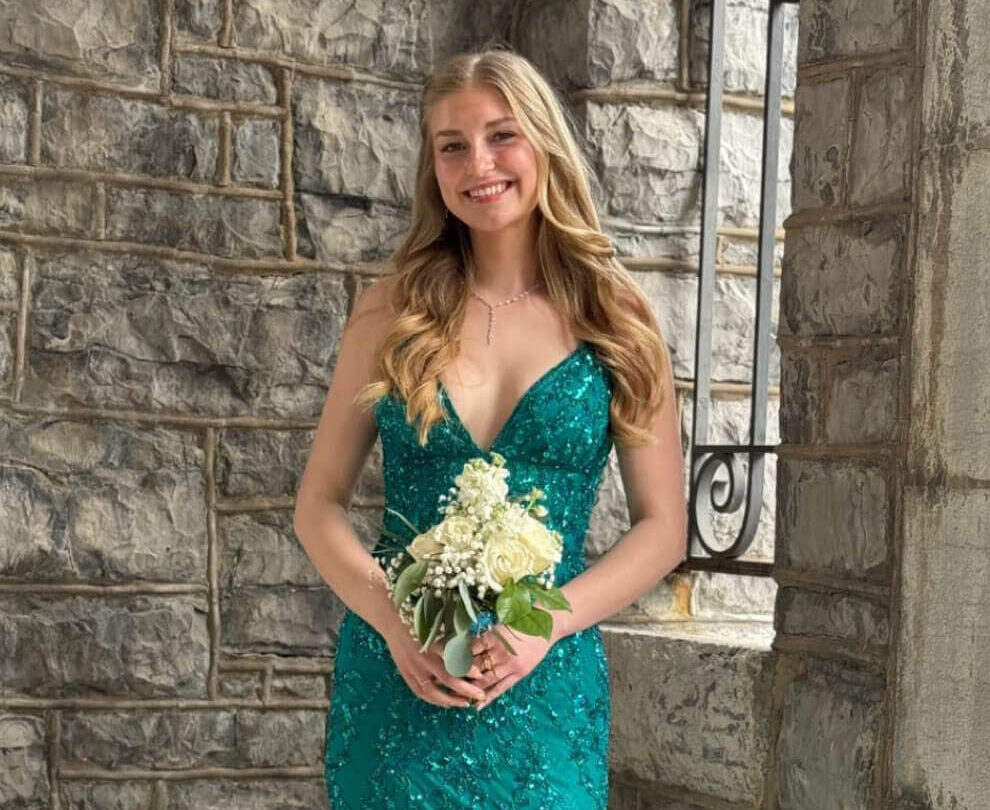 A woman poses in front of a stone wall wearing a fitted emerald green floor-length gown with shimmering embellishments while holding a white floral bouquet.