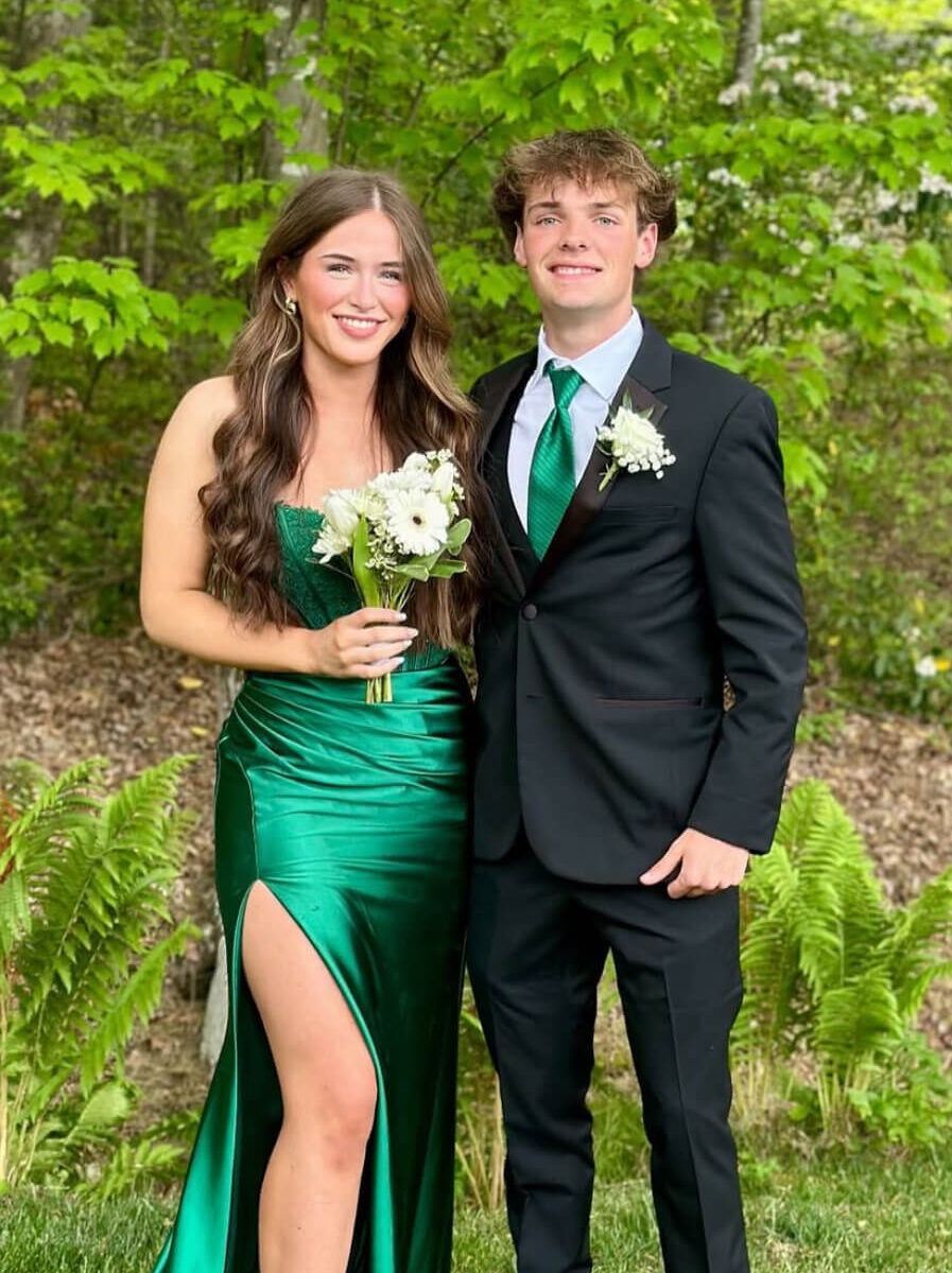 A couple poses outdoors, with the woman wearing an emerald green strapless satin gown with a high slit and holding white flowers and the man wearing a black suit with a green tie.