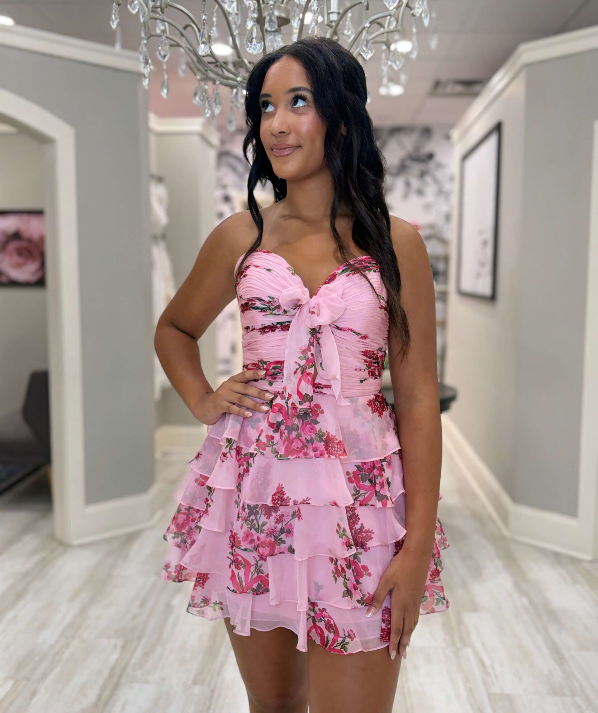 A woman poses indoors beneath a chandelier wearing a short pink floral dress with a sweetheart neckline, bow detail, and tiered ruffle skirt.