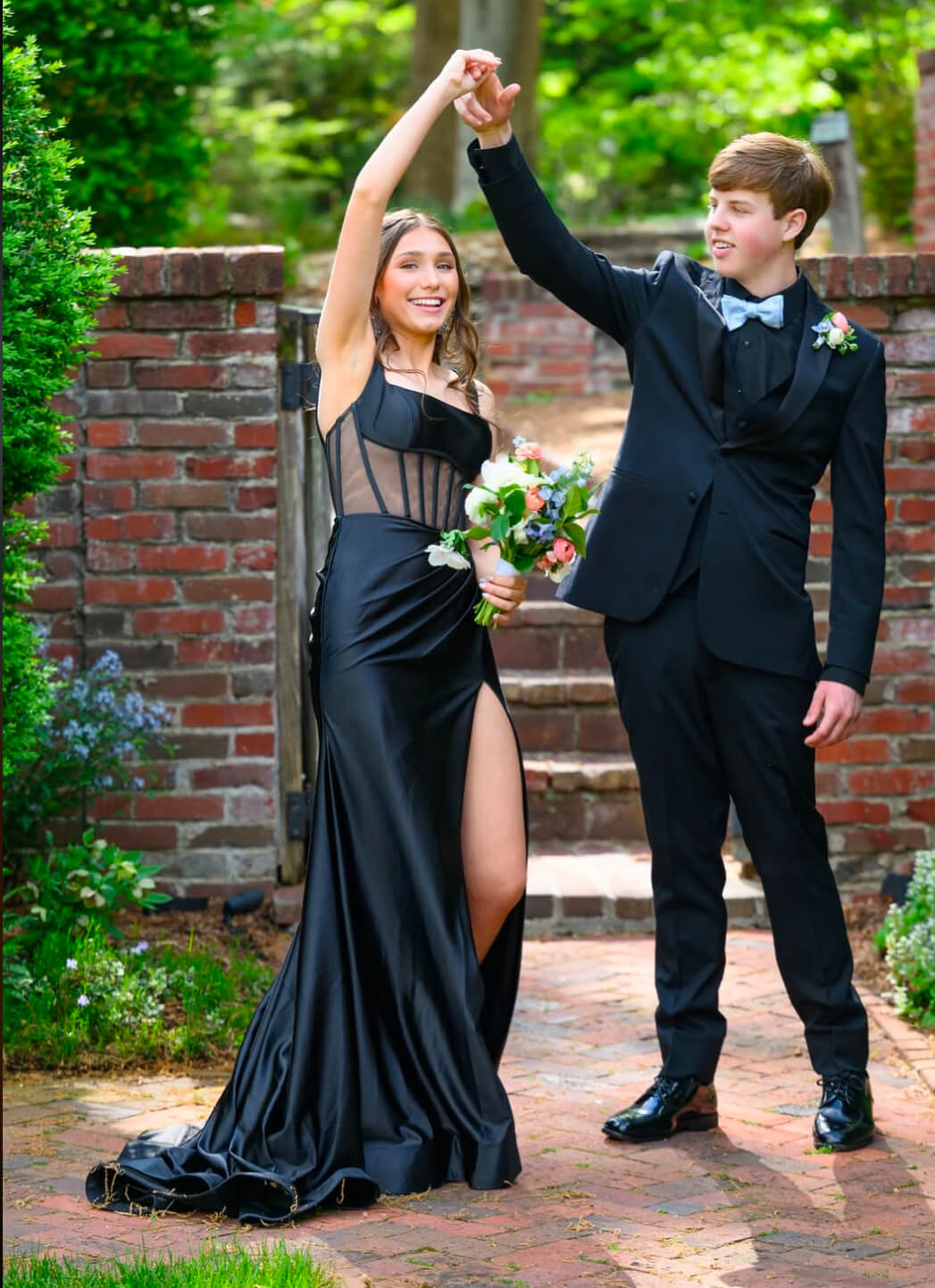 A couple dances outdoors on a brick walkway, with the woman wearing a black floor-length gown with a sheer corset bodice and high slit and the man wearing a black suit with a silver bow tie.