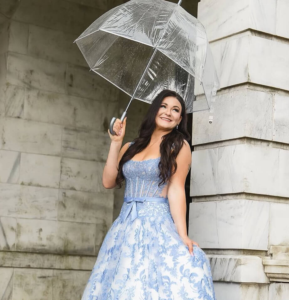 A woman stands beneath a clear umbrella wearing a blue-and-white patterned ball gown with a corset-style bodice and full skirt.