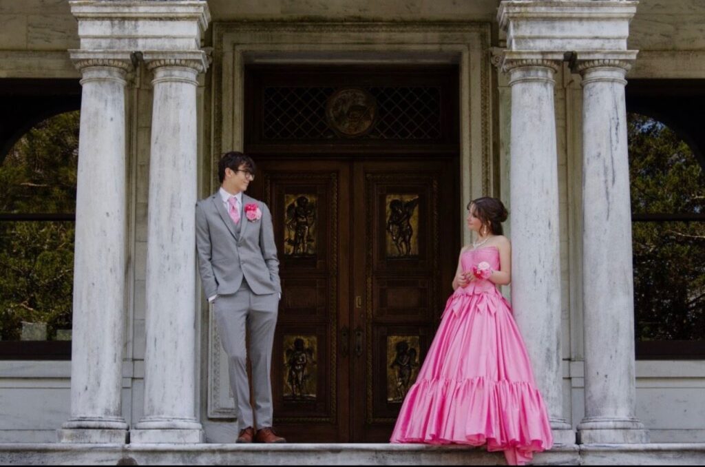 A couple poses between large columns in front of ornate wooden doors, with the woman wearing a bright pink strapless ball gown and the man wearing a gray suit with pink accessories.