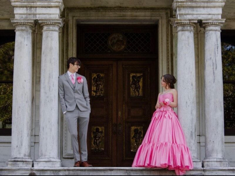 A couple poses between large columns in front of ornate wooden doors, with the woman wearing a bright pink strapless ball gown and the man wearing a gray suit with pink accessories.